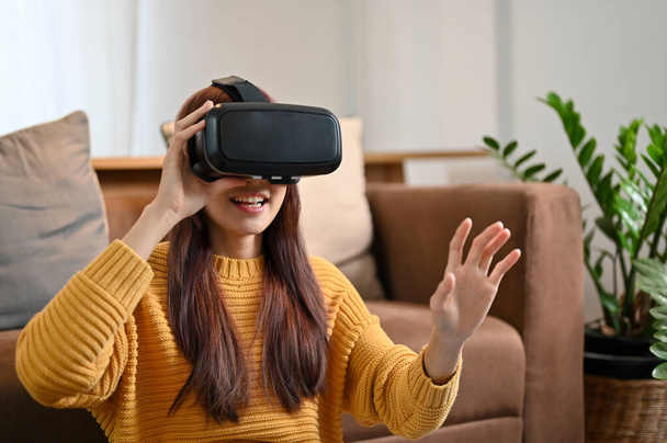 Femme asiatique millénaire excitée jouant à un jeu de virtualité avec des lunettes VR dans le salon moderne. VR gaming, casque VR. - Photo, image