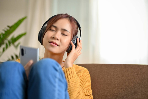 Relaxed millennial Asian woman on the sofa in the living room, listening to music with wireless headphones. - Foto, imagen