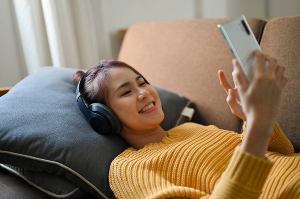 Schöne asiatische Millennial-Frau auf bequemem Sofa liegend, Musik hörend, mit drahtlosen Kopfhörern und Smartphone. - Foto, Bild