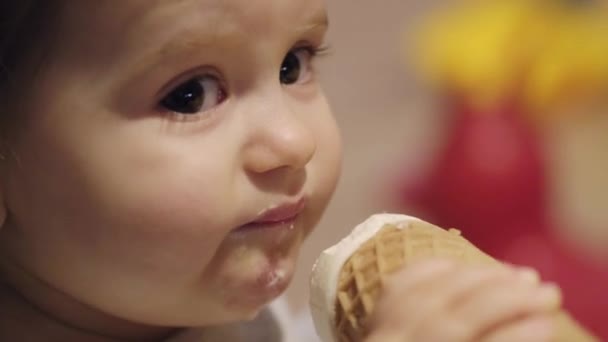 Retrato de cerca de una linda niña lamiendo helado de vainilla en cono, sentada en casa. Bebé lamiendo comida congelada. Feliz estilo de vida infantil. Sabroso verano - Imágenes, Vídeo