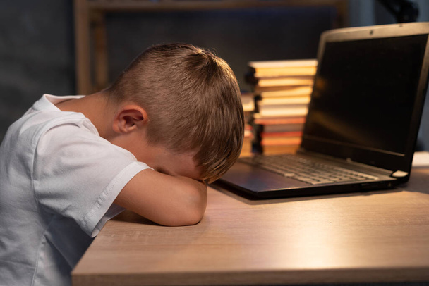 Le gamin fatigué s'est endormi à côté d'un ordinateur portable faisant des devoirs scolaires la nuit, gros plan. Étudiant garçon épuisé dormant sur le bureau devant l'ordinateur, concept de retour à l'école - Photo, image
