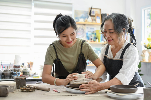 Profesionální Aged žena učitelka učí hezkou mladou ženu, aby keramické misky s hlínou v dílně. tvůrčí činnost a koncepce koníčků. - Fotografie, Obrázek