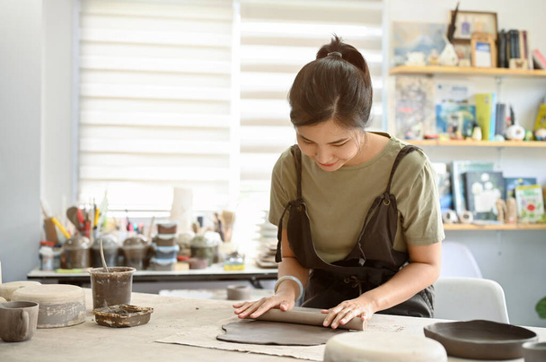 Relaxado e calmo millennial asiático mulher rolando argila na mesa de trabalho com rolo de argila na oficina de artesanato. Conceito de cerâmica de moldagem - Foto, Imagem