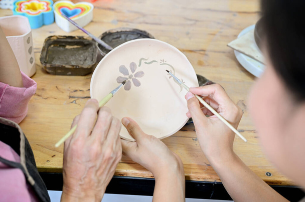 Nahaufnahme, Zwei asiatische Frauen beim gemeinsamen Bemalen einer schönen Blume in einem Keramikteller mit Acrylfarbe in der Werkstatt. - Foto, Bild