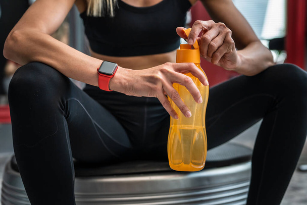Midsection of unknown caucasian woman female athlete holding plastic bottle of water while taking a brake resting during training sitting on the power plate vibration platform copy space - Photo, Image