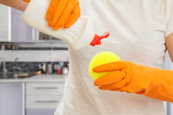 Жінка в футболці тримає жовту губку і пластикову пляшку пральної рідини на кухні. Концепція миття та очищення
. - Фото, зображення