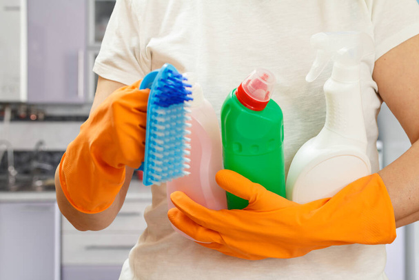 Γυναίκα σε ένα λευκό t-shirt και γάντια από καουτσούκ κρατώντας πλαστικά μπουκάλια υγρού πλύσης και ένα πινέλο στην κουζίνα. Έννοια πλύσης και καθαρισμού. - Φωτογραφία, εικόνα
