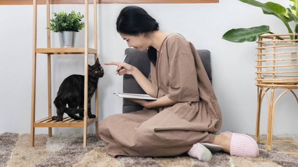 アジアの女性は喜んで日記を書き、リビングルームで猫と遊んで座っています,最愛のペット,親友ペット,ハッピーエリア,自宅での休日,リラックスして楽しみを持っています,猫とカーペットの上に座って. - 写真・画像