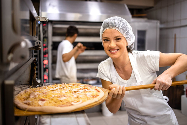 Portret kobiety piekarza trzymającej pyszną pizzę przy piekarniku w restauracji pizzeria. - Zdjęcie, obraz
