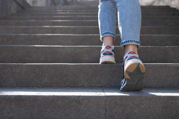 Großaufnahme einer Frau in Jeans und Turnschuhen, die eine steile Treppe hinaufsteigt. Touristin in legerer Kleidung geht nach draußen - Foto, Bild