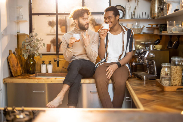 Two guys of different ethnicity having warm conversation while sitting with coffee on kitchen at home. Concept of close male friendship or relationship as gay. Caucasian and hispanic man together - Foto, immagini