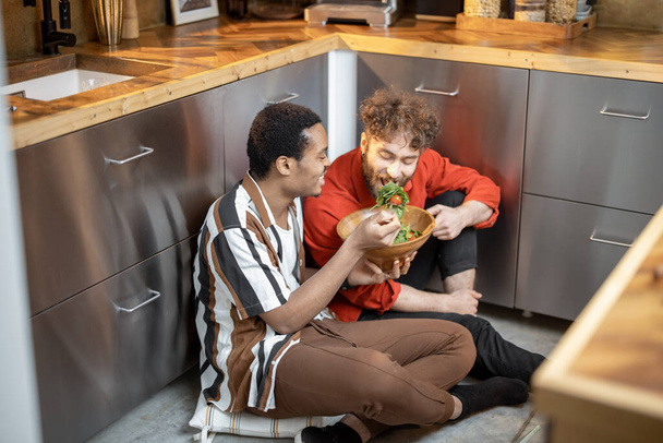 Two brightly dressed stylish guys having close conversation, sitting together on a kitchen floor and eating healthy salad. Concept of gay relations. Caucasian and latin man bonding while talking close - Фото, изображение