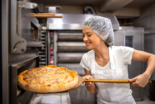 Pizza master taking freshly baked crunchy pizza from the oven in bakery. - Photo, Image
