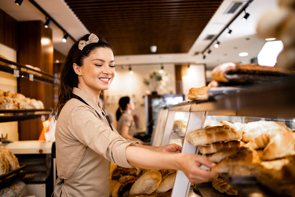 Piękna pracownica delikatesów w mundurze sprzedająca świeże wypieki i hodowana w dziale piekarniczym supermarketu. - Zdjęcie, obraz