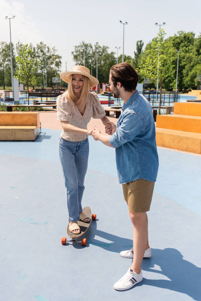 Young man holding hands of girlfriend in sun hat riding longboard in skate park  - Foto, Bild