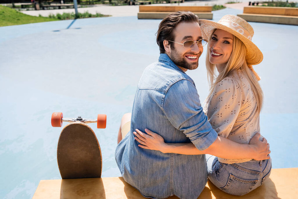 Positive couple hugging and looking at camera near longboard in skate park  - Φωτογραφία, εικόνα
