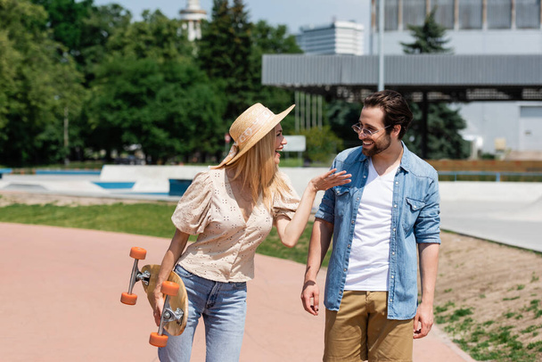 Cheerful woman in sun hat holding longboard and talking to boyfriend in skate park  - Photo, image