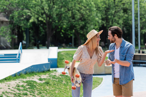 Vista lateral de pareja positiva con longboard hablando en skate park  - Foto, Imagen