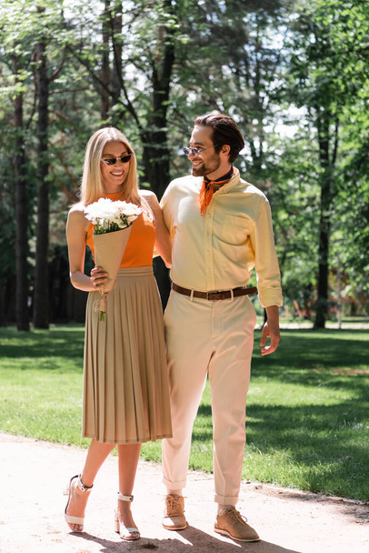 Stylish woman holding bouquet near smiling boyfriend with sunglasses in park  - Фото, изображение