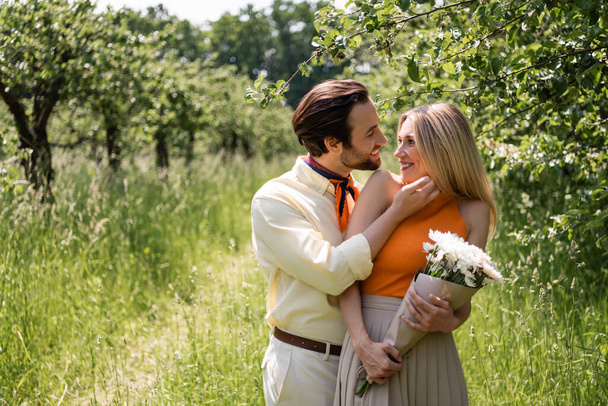 Cheerful man touching stylish girlfriend with bouquet in park  - Fotografie, Obrázek