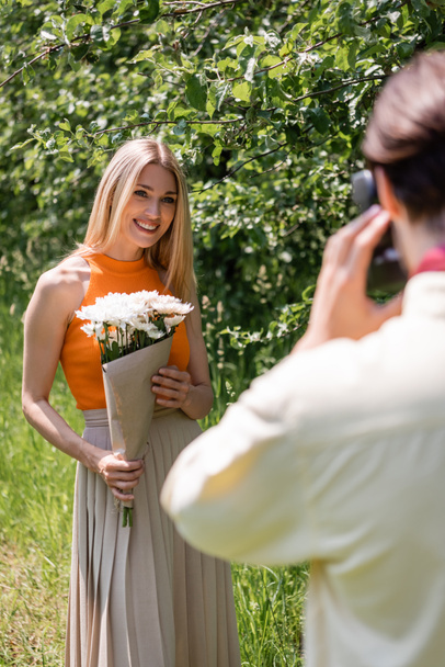 Femme souriante tenant bouquet près de petit ami flou avec caméra vintage dans le parc  - Photo, image