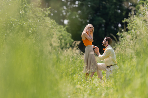 Side view of stylish man holding hand of girlfriend while kneeling in summer park  - Φωτογραφία, εικόνα