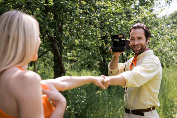 Homme à la mode tenant caméra rétro et tenant la main de petite amie floue dans le parc  - Photo, image