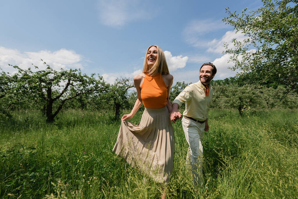 Cheerful woman holding hand of boyfriend on lawn in park  - Zdjęcie, obraz