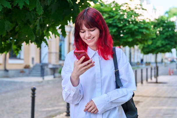 Beautiful smiling teenage girl looking at phone on city street. Female teenager with red dyed hair with backpack using smartphone. Lifestyle, technology, communication, youth, young people - Фото, зображення