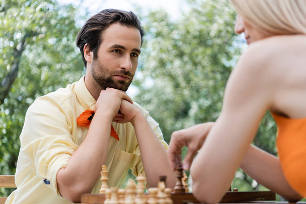 Stylish man looking at blurred girlfriend playing chess in park  - Fotó, kép