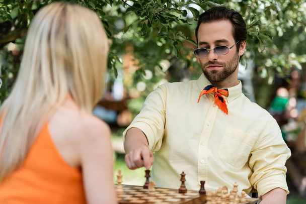 Trendy νεαρός άνδρας που δείχνει σκακιστική φιγούρα κοντά θολή φίλη στο καλοκαιρινό πάρκο  - Φωτογραφία, εικόνα