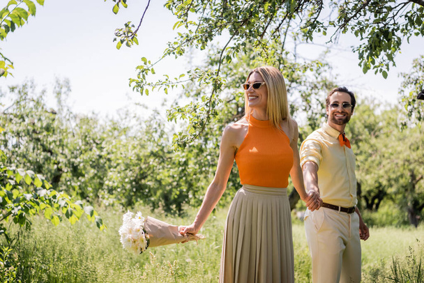 Happy woman holding bouquet and hand of trendy boyfriend in summer park  - Zdjęcie, obraz