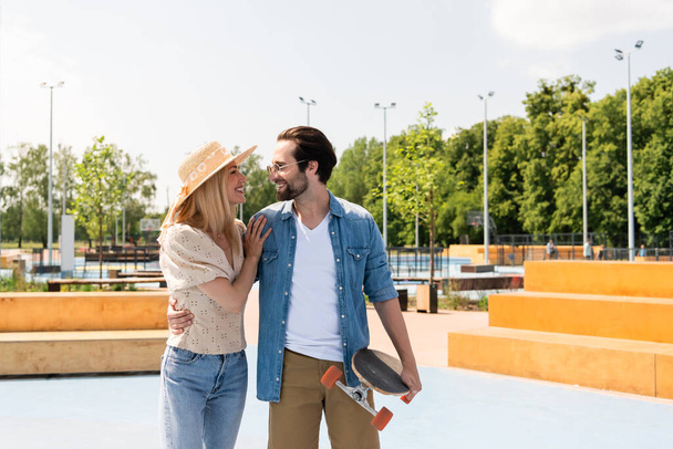Vista laterale della donna sorridente che abbraccia il giovane fidanzato con longboard nello skate park  - Foto, immagini