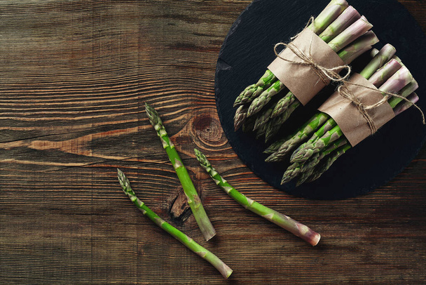 Bunches of an edible, mouth-watering stalks of asparagus on a stone slate, wooden background. Fresh, green vegetables, top view. Healthy meal. Fall harvest, agricultural farming concept. - Foto, afbeelding