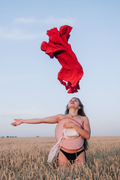 A young, slender girl with loose hair in a field of wheat hides behind a hat and tosses a red dress. High quality photo - Foto, imagen