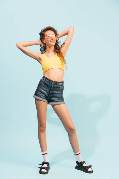 Portrait of smiling young girl with curly hair posing in casual comfortable summer outfit isolated over light blue studio background. Concept of youth, beauty, lifestyle, fashion, fun, emotions - Foto, afbeelding