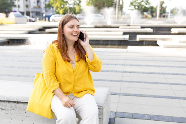 A portrait of a smiling beautiful woman talking on phone on urban background. Happy woman with yellow shopping bag is using a smartphone outdoors, spring time. Traveler - Zdjęcie, obraz