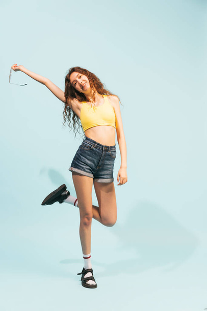 Portrait of cheerful young girl in jeans shorts, yellow top posing isolated over light blue background. Summertime mood, Concept of youth, beauty, lifestyle, fashion, fun, emotions, party - Photo, image
