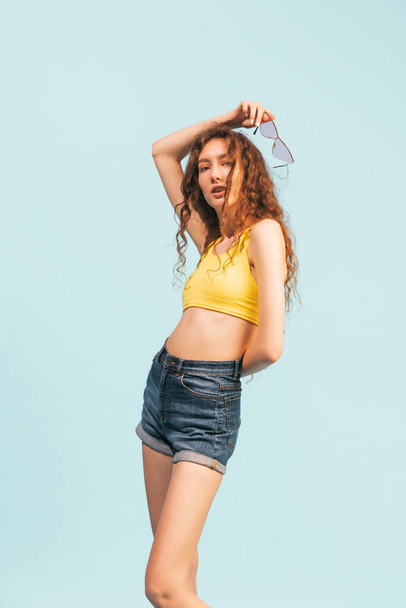 Portrait of young girl with long gurly hair posing in casual summer outfit isolated over light blue studio background. Concept of youth, beauty, lifestyle, fashion, fun, emotions, style - Photo, Image