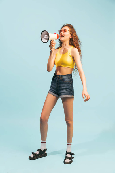 Portrait of young girl in summertime outfit, shorts and top, posing, shouting in megaphone isolated over light blue studio background. Concept of youth, beauty, lifestyle, fashion, fun, emotions - Photo, Image
