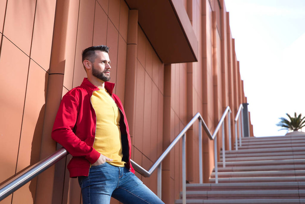 Joven guapo con chaqueta roja, camiseta amarilla y jeans apoyados en la barandilla de una escalera. Concepto belleza, moda, moderno, de moda. - Foto, imagen