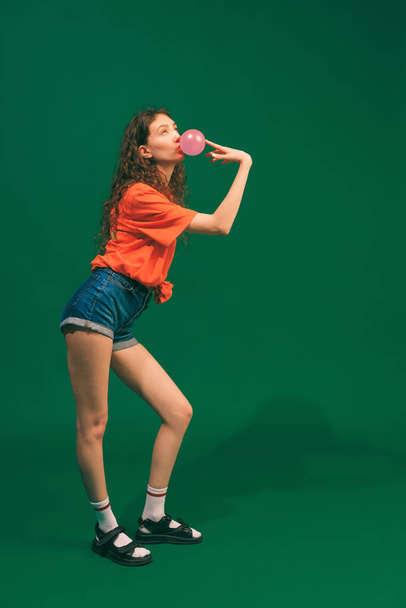 Full-length portrait of young girl posing in jeans shorts, orange T-shirt and sandals, eating bubble gum isolated over green background. Concept of youth, beauty, lifestyle, fashion, fun, emotions - Foto, afbeelding