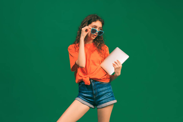 Портрет молодой девушки, студентки в повседневной одежде и солнцезащитных очках с планшетом, позирующим изолированно на зеленом фоне студии. Онлайн-редактирование. Молодость, красота, образ жизни, мода, веселье, эмоции - Фото, изображение