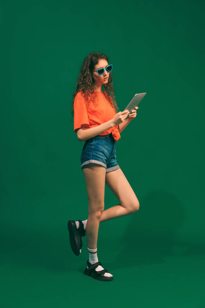 Portrait of young girl in casual outfit, shorts and T-shirt, holding tablet, posing isolated over green studio background. Education. Concept of youth, beauty, lifestyle, fashion, fun, emotions - Photo, image