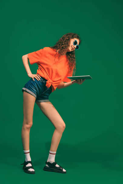 Retrato de chica joven en traje casual, pantalones cortos y camiseta, sosteniendo la tableta, posando aislado sobre fondo de estudio verde. Servicios en línea. Concepto de juventud, belleza, estilo de vida, moda, diversión, emociones - Foto, Imagen