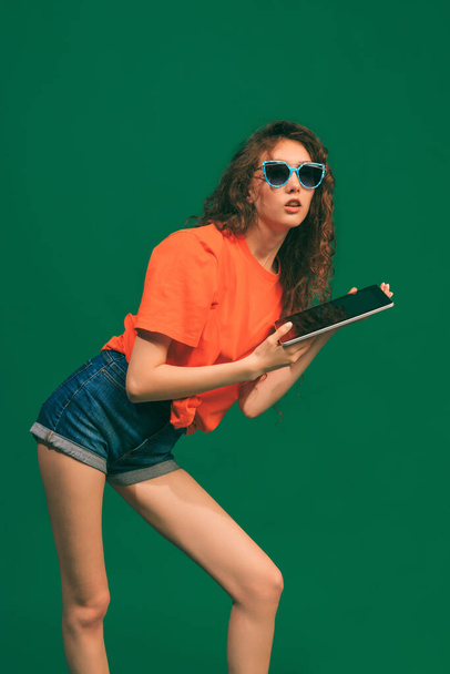 Portrait of young girl in casual outfit, shorts and T-shirt, holding tablet, posing isolated over green studio background. Concept of youth, beauty, lifestyle, fashion, fun, emotions - Photo, Image