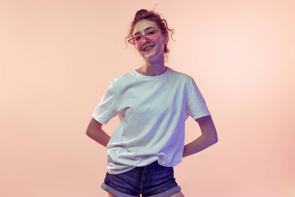 Retrato de una joven elegante con gafas de sol de moda, posando en aislado sobre fondo de estudio rosa en luz de neón. Parece feliz. Concepto de juventud, belleza, estilo de vida, moda, diversión, emociones - Foto, imagen