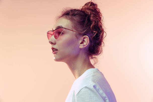 Portrait of stylish young girl in trendy sunglasses, posing in white T-shirt isolated over pink studio background in neon light. Side view. Concept of youth, beauty, lifestyle, fashion, fun, emotions - Photo, Image