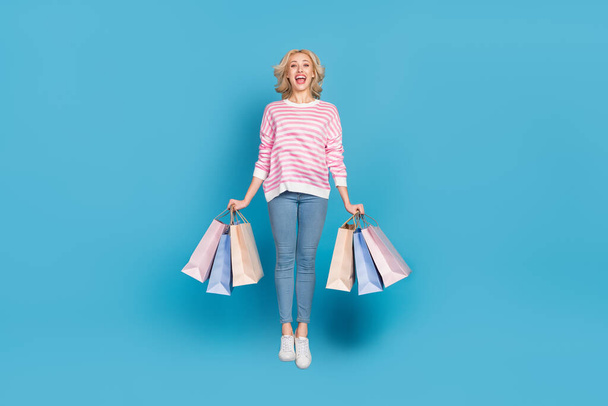 Photo of shiny excited woman wear striped sweater jumping high holding bargains isolated blue color background. - Foto, afbeelding