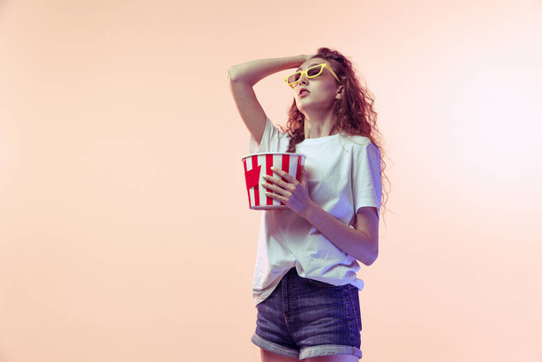 Ritratto di giovane ragazza emotiva in abito casual in posa con cesto di popcorn isolato su sfondo rosa studio in luce al neon. Concetto di gioventù, bellezza, stile di vita, moda, divertimento, emozioni - Foto, immagini